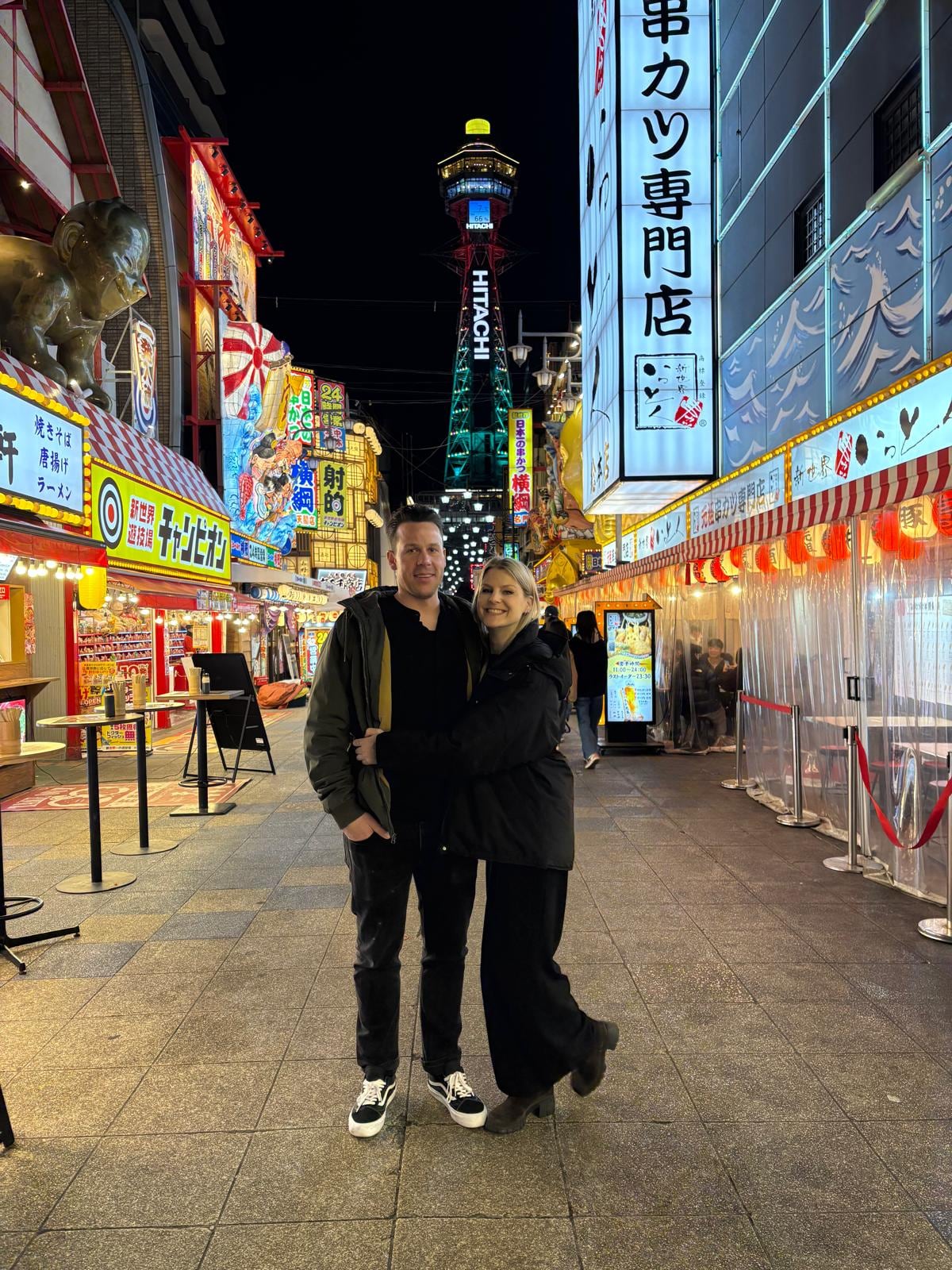 Robin Heer und Melanie Meier nachts in Osaka, im Quartier Shinsekai, auf ihrer Reise durch Japan. Im Hintergrund der beleuchtete Tsutenkaku-Turm.